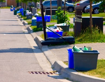 A street scene in Gunnersbury showing a variety of rubbish accumulation along the pavement, including several blue and grey plastic bins, some with lids open revealing discarded items such as paper and plastic containers. There are also cardboard boxes and loose rubbish piled next to the bins, with some items spilling onto the curbside. A broom and dustpan rest against a black bin trolley, suggesting ongoing cleaning activity. The background features parked cars, small trees, and greenery, indicating a residential area. The pavement is adjacent to a grassy verge bordered by flower beds, with sunlight casting shadows across the scene. This image illustrates the need for professional waste removal services, such as those provided by Commercial Waste Removal Gunnersbury, to efficiently clear rubbish from residential streets in West London postcodes, supporting cleanliness and environmental standards in the local community.