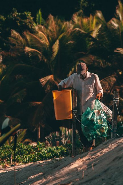 A man wearing a beige cap, glasses, and a light-colored long-sleeve shirt is walking outdoors during the daytime, carrying a green plastic bag filled with waste in his left hand and holding an orange 